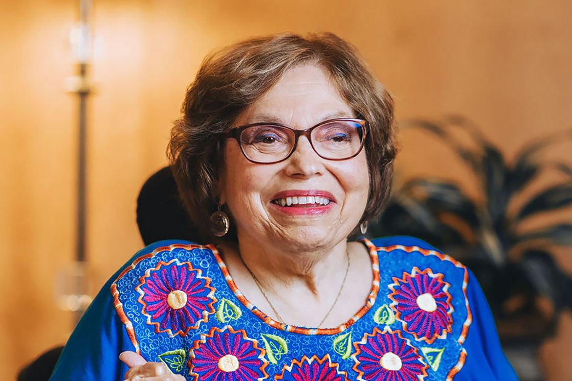 Judy, an older woman with light skin, wears glasses and a vibrant floral shirt. She smiles.