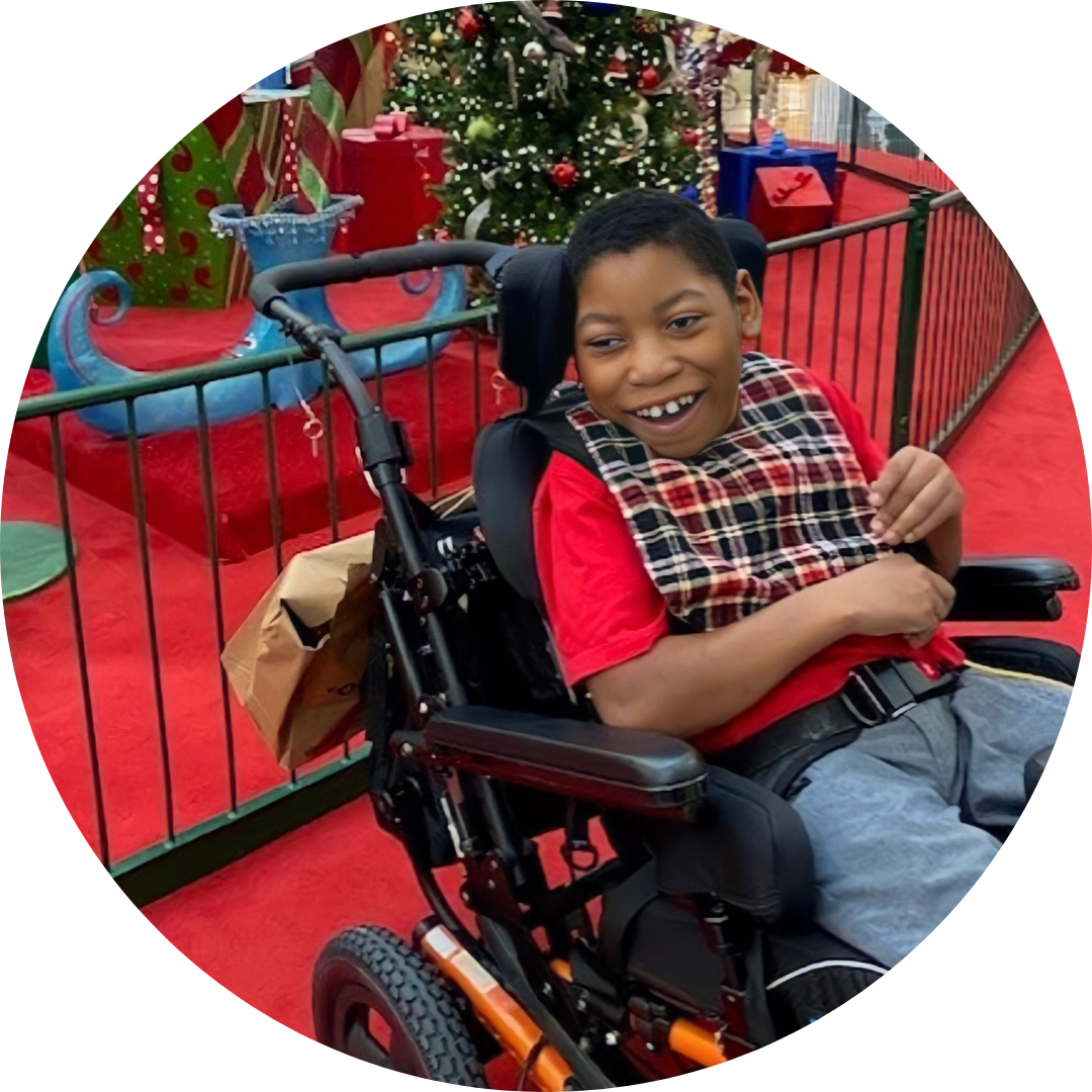Lorenzo, a young boy with brown skin, short black hair, wearing a red shirt and blue shorts, seated in a wheelchair in front of Christmas tree display