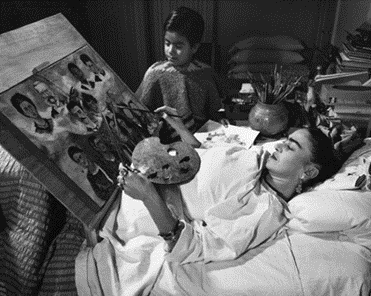 Frida Kahlo laying down in bed painting using a bed accessible easel and paints