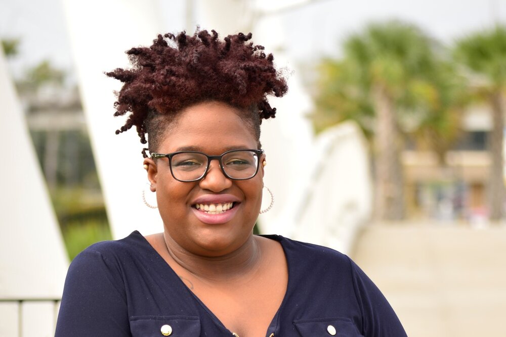 Sherona Bryant, a woman with dark skin and short reddish brown hair twists wears glasses and a v neck blue shirt. 