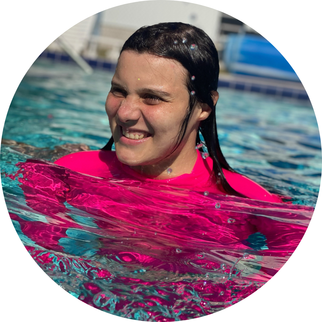 Ariana, a young woman with light skin, dark brown hair, wearing a bring pink shirt smiles wild standing in a swimming pool.