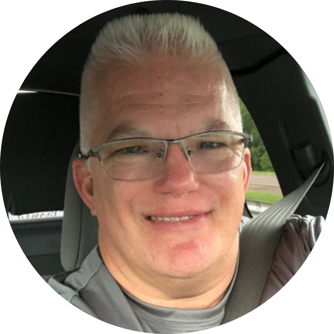  image of Calvin in his car. He has light skin, blonde short hair, wears glasses and a grey tshirt and smiles. 