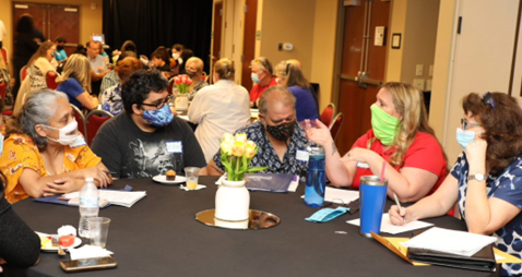 5 individuals seated at a table at a Public Forum. They wear face masks and have informational papers laid out on the table.