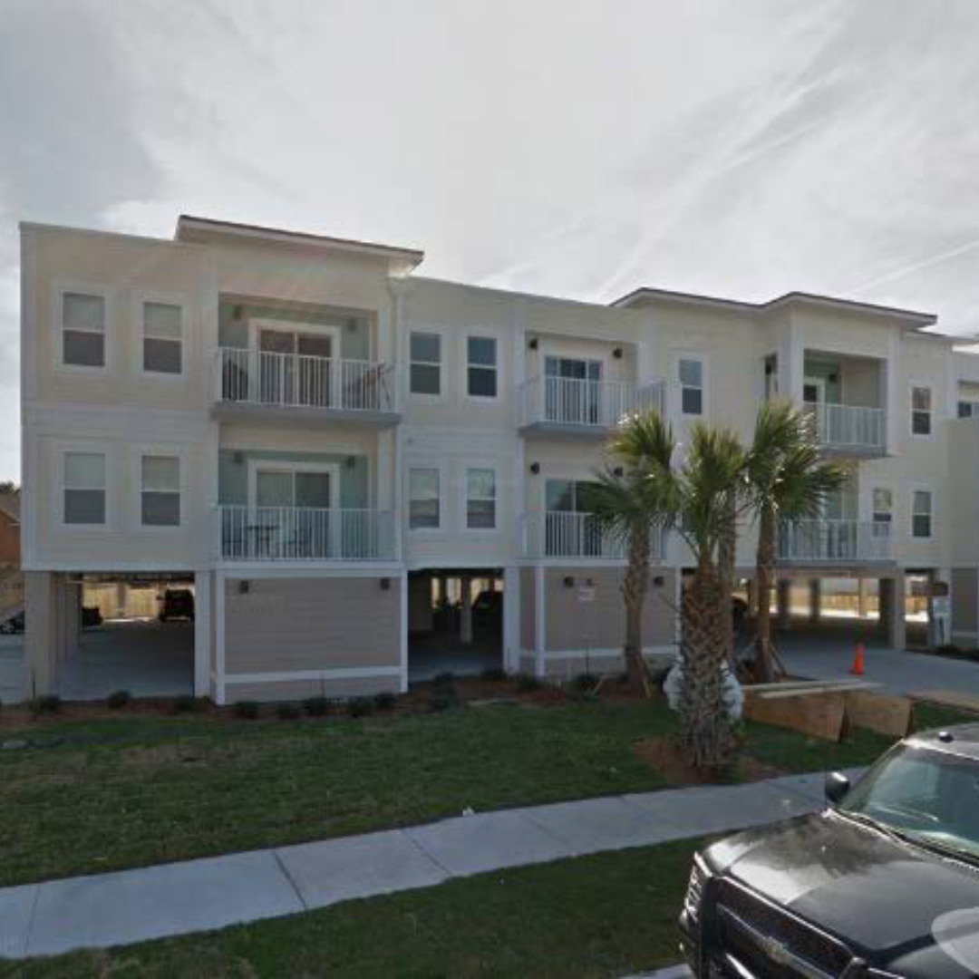 A 3-story beige and tan apartment complex surrounded by palm trees and grass.