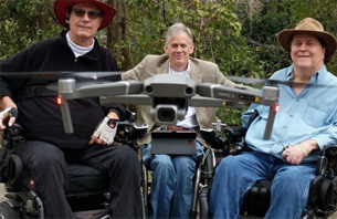 3 men using wheelchairs posing together for a photo. A drone hovers in front of them, which is a tool they use to take photos.
